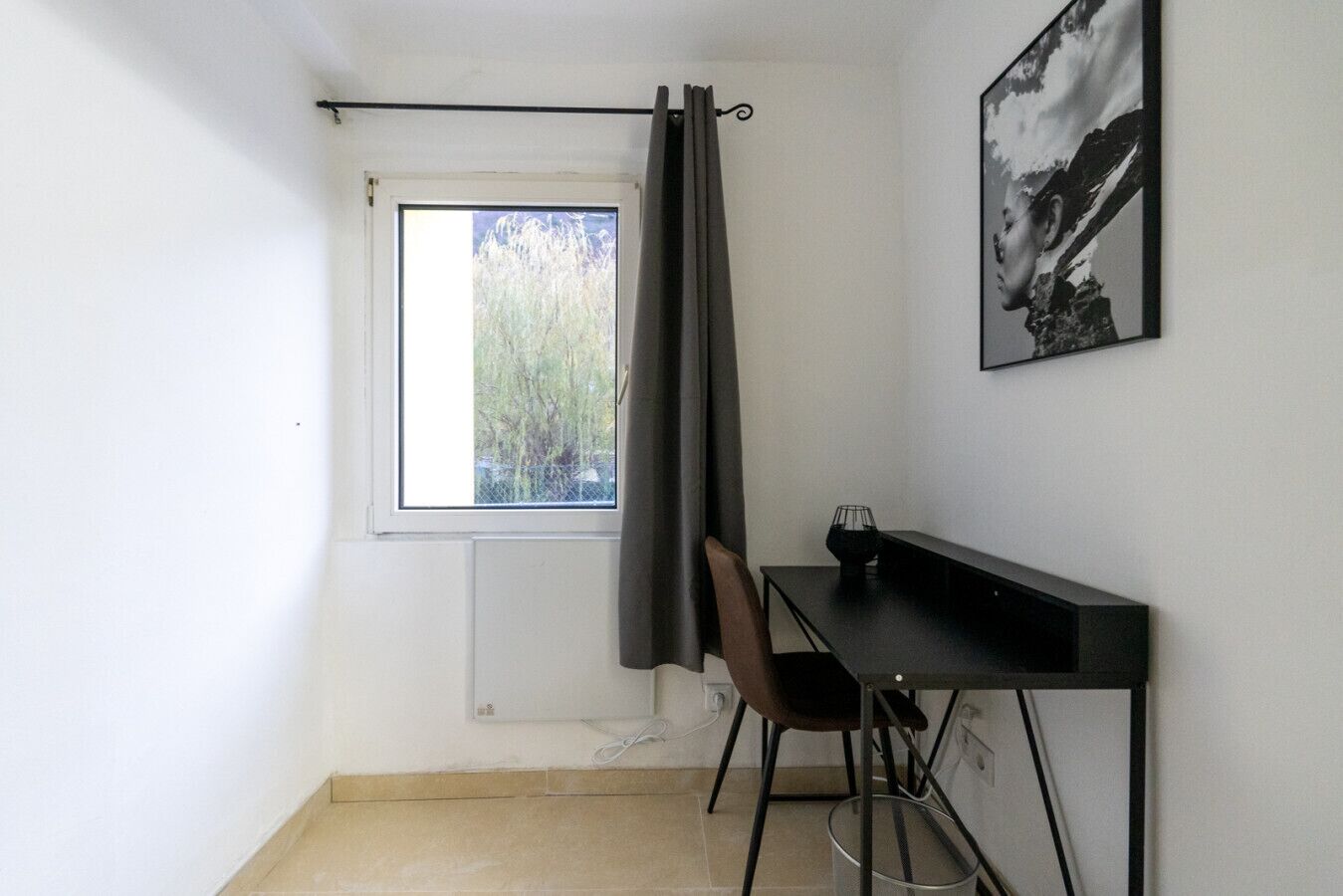 Una habitació petita amb una finestra, un escriptori, una cadira i un quadre al mur.