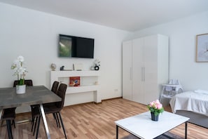 A modern room with a dining table, chairs, a TV, a wardrobe, a bed, and a small table with flowers. - 55 m² I 2 Rooms I Up to 5 Guests I Near Vienna (Kaltenleutgeben)