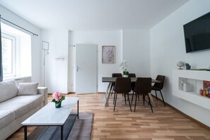 A modern living room with a sofa, a coffee table with flowers, a dining area with chairs, and a TV mounted on the wall. - 55 m² I 2 Rooms I Up to 5 Guests I Near Vienna (Kaltenleutgeben)