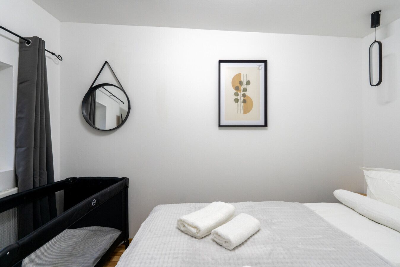 Ein Schlafzimmer mit einem Babybett, einem Bett mit weißer Bettwäsche, einem gerahmten abstrakten Kunstwerk, einem hängenden Spiegel und einem Vorhang.