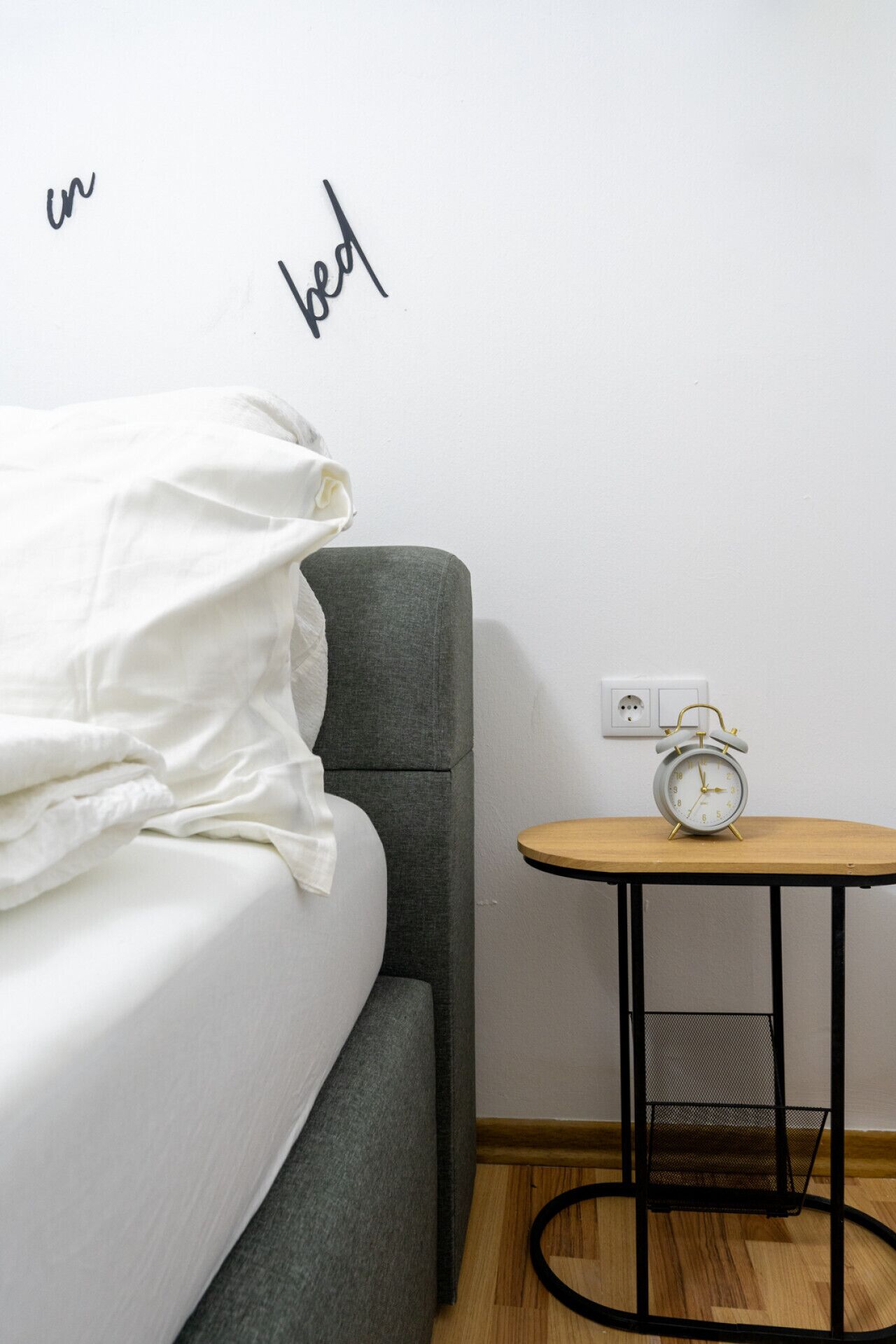 Ein ordentlich bezogenes Bett mit weißen Leinen, ein grau gepolsterter Sessel, ein kleiner runder Holztisch mit schwarzem Metallgestell und eine weiße Wand mit Steckdose.