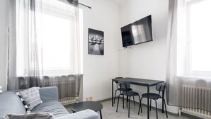 A modern living room with a grey sofa, a small round table, a dining table with four chairs, a flat-screen TV mounted on the wall, and a radiator. - Stylish Apartment at Main Square - 35 m² - Quiet (Leoben)