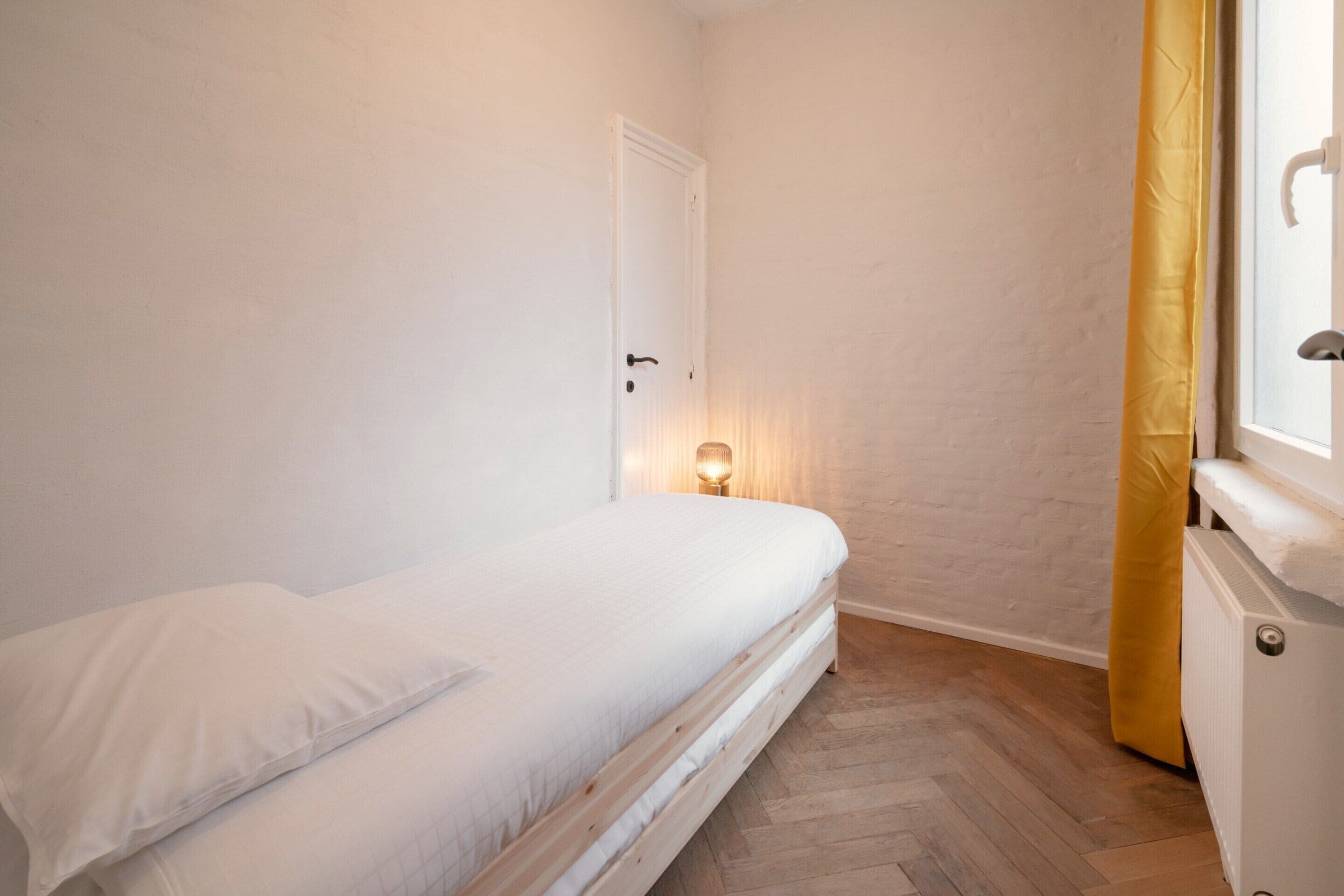 Une chambre à coucher avec un lit, une porte, un radiateur et une fenêtre avec des rideaux.
