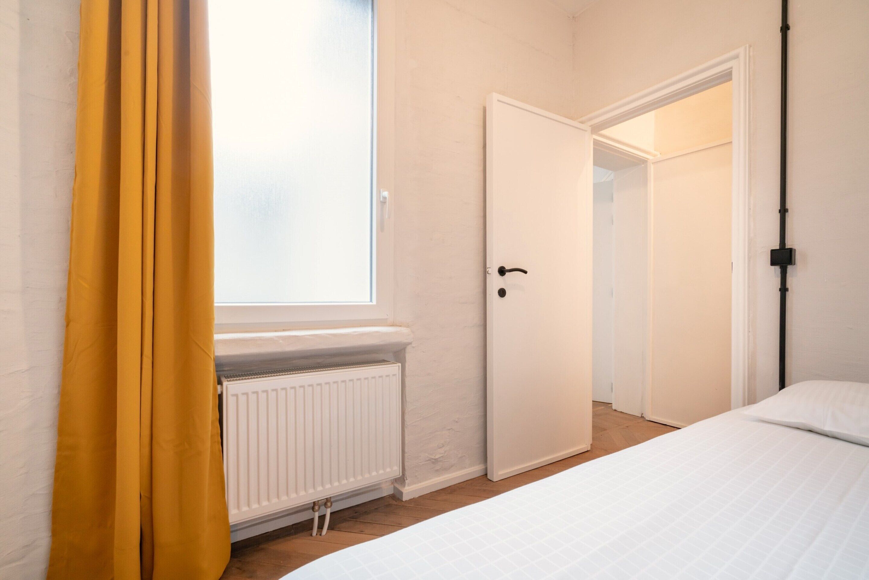 Un dormitorio con cama, un radiador, una ventana con cortinas y una puerta.