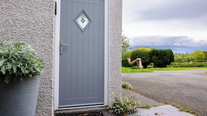 A grey door with a diamond-shaped window, a potted plant, and a view of a green landscape with a tree and a fence. - Eryri View Retreat (Beaumaris)