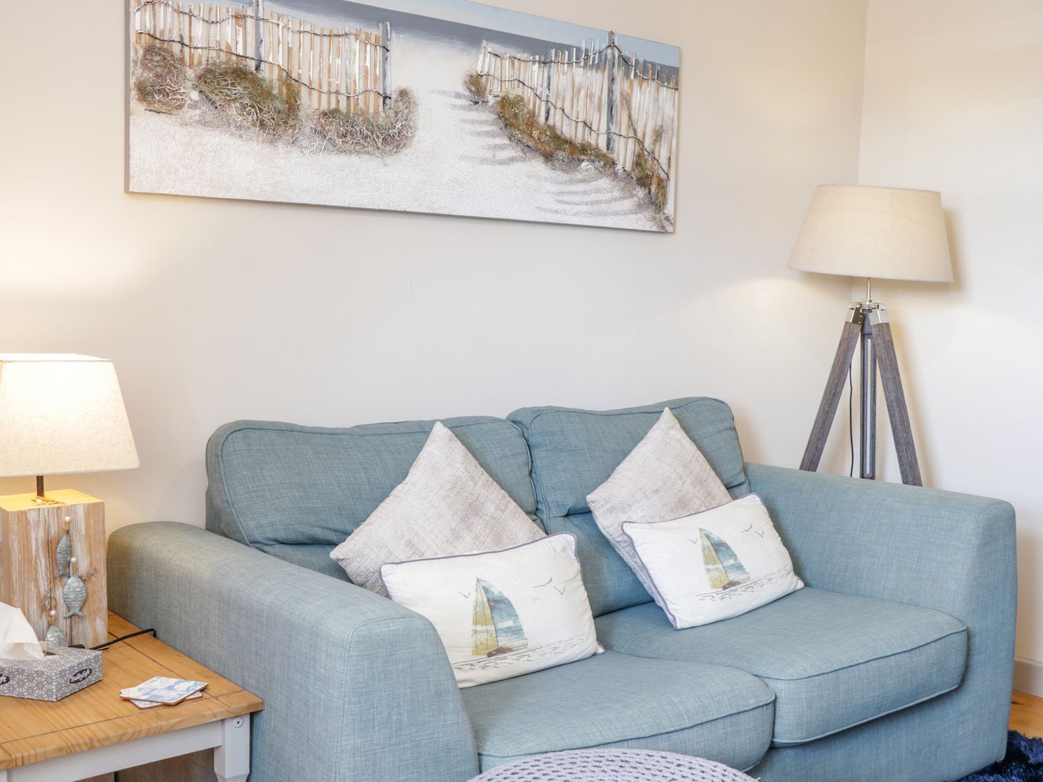 A room with a light blue sofa, two decorative pillows, a wooden side table with a lamp, a floor lamp, and a painting of a beach scene.