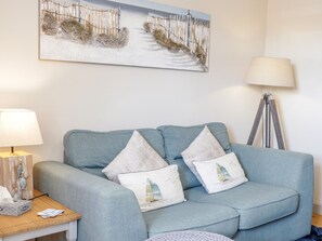 A room with a light blue sofa, two decorative pillows, a wooden side table with a lamp, a floor lamp, and a painting of a beach scene.