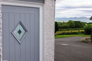 A grey door with a diamond-shaped window, a white textured wall, and a view of a rural landscape with fields and mountains.