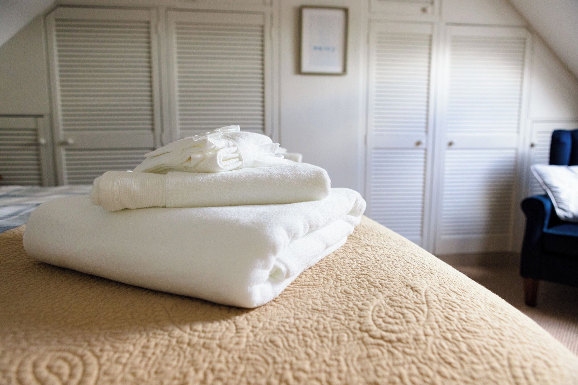 Una pila ordenada de toallas blancas sobre una cama color beige con textura, en una habitación con armarios blancos y un cuadro en la pared.
