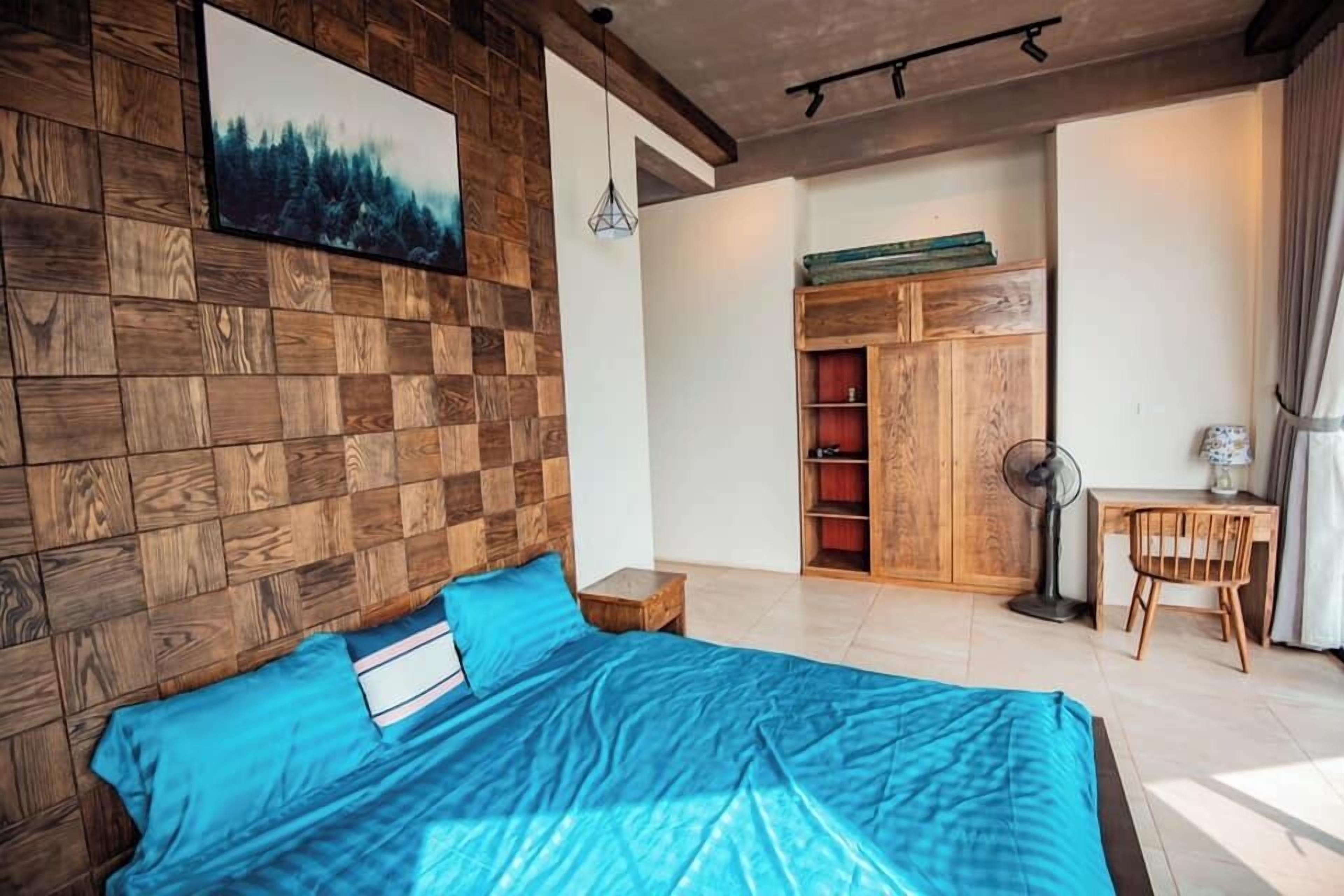A bedroom with a wooden headboard, a bed with blue bedding, a desk with a chair, a fan, and a wardrobe.