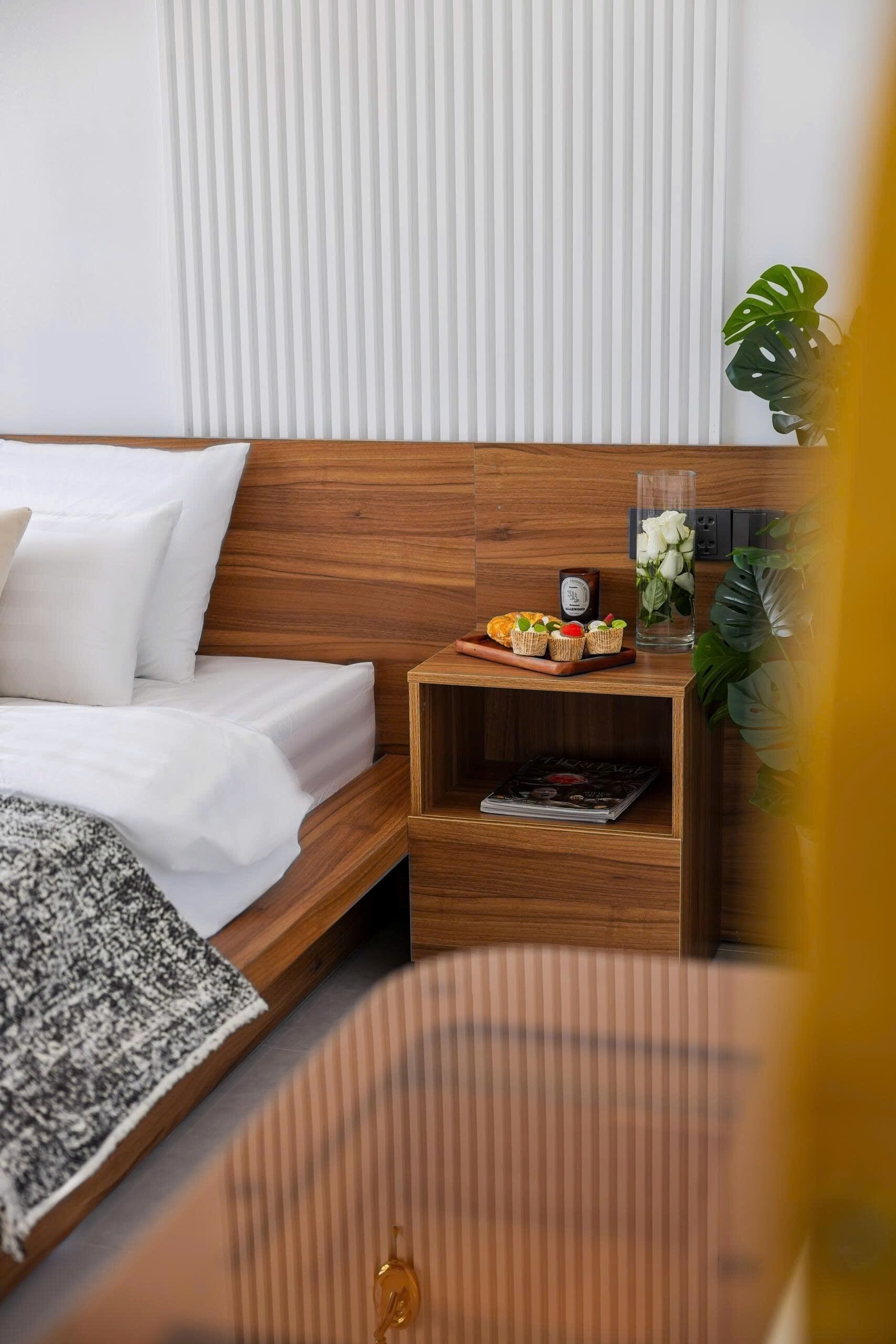 A hotel room with a wooden headboard, a bed with white linens, a nightstand with a plant, a vase with flowers, and a magazine on the nightstand.