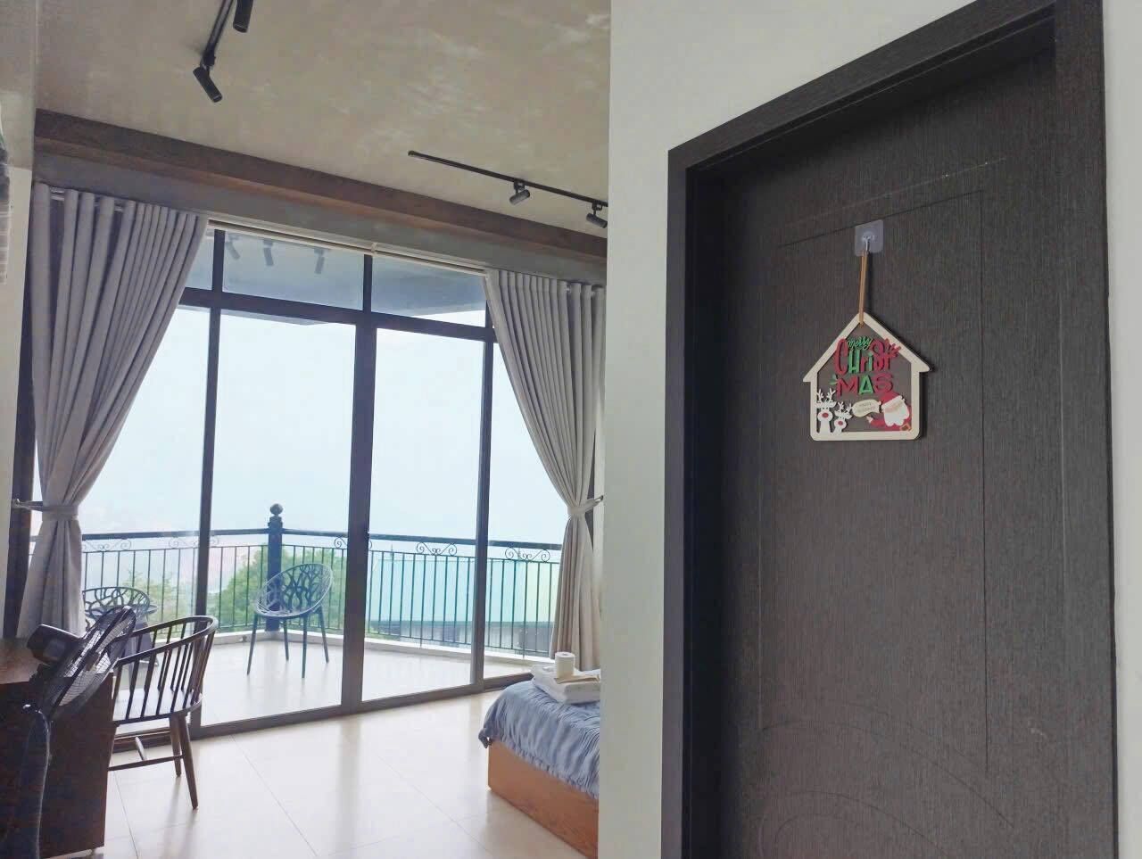Una habitación con un amplio ventanal corredero que da a un balcón, una cama, un escritorio con una silla y un adorno colgado en la puerta.