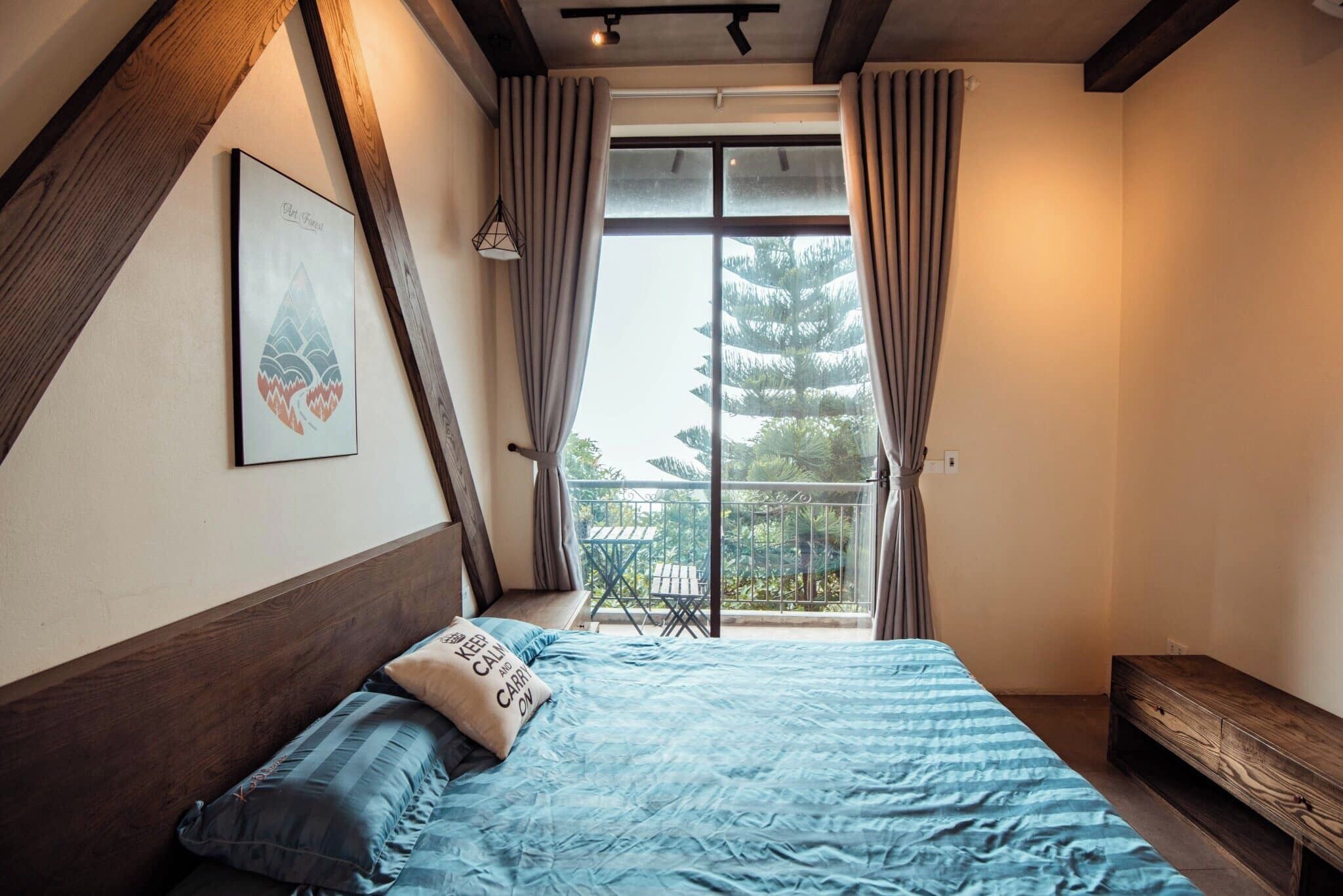 A bedroom with a bed, a bench, a picture on the wall, and a balcony with a view of trees.