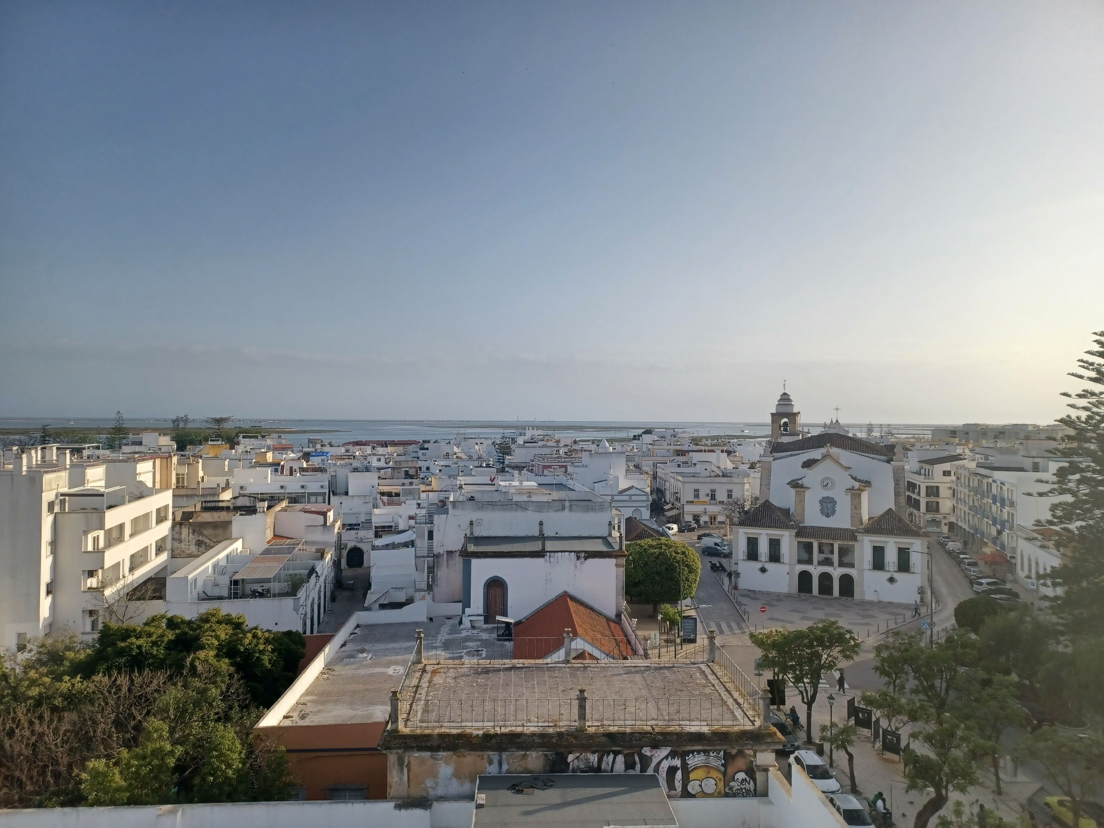 A cityscape with white buildings, a church with a bell tower, and a view of the sea.