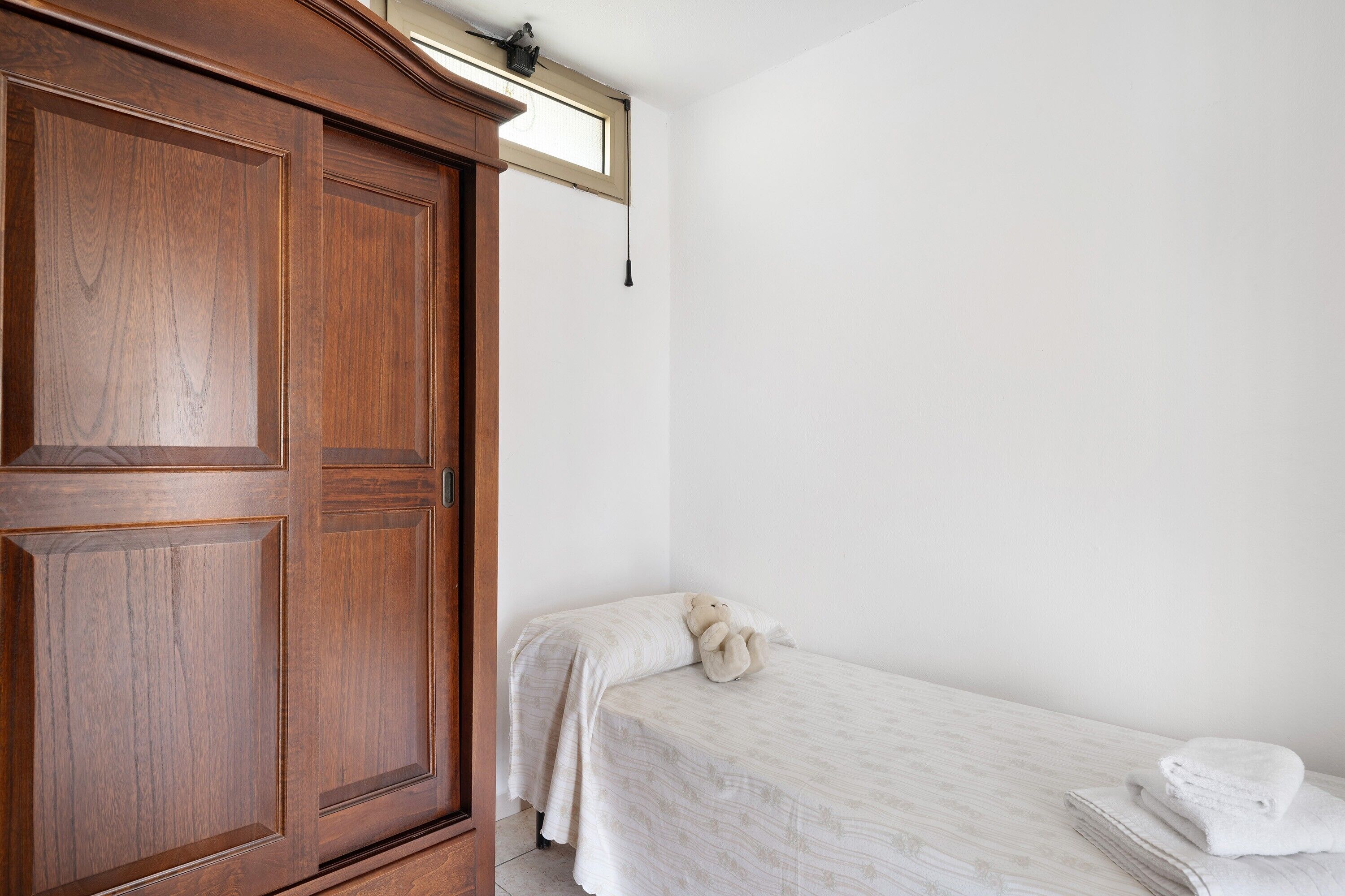 Ein Schlafzimmer mit Holzschrank, einem Bett mit weißer Bettdecke, einem Teddybären und einem Fenster mit Pendelleuchte.