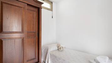 Ein Schlafzimmer mit Holzschrank, einem Bett mit weißer Bettdecke, einem Teddybären und einem Fenster mit Pendelleuchte.
