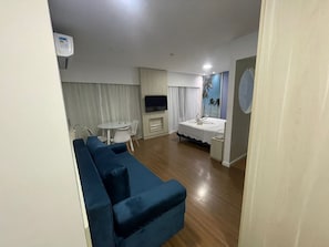 A living room with a blue sofa, a small round table, a TV, and a bed with a white bedspread.