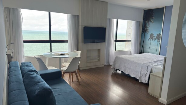 Ein modernes Hotelzimmer mit einer Couch, einem kleinen Tisch, einem Bett, einem Fernseher und großen Fenstern mit Blick aufs Meer.