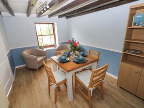 Een eetgedeelte met een houten tafel voor vier personen, omringd door stoelen met kussens. Er is een raam met uitzicht op groen, en een boekenkast met boeken en decoratieve objecten.