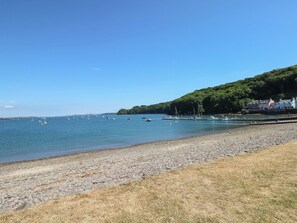 A pebbly beach with boats, a clear sky, and a forested hillside. - Beacon House (Haverfordwest)
