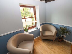 A room with a curved beige armchair, a smaller armchair, a window with a view of greenery, a lamp on a windowsill, and a potted plant. - Beacon House (Haverfordwest)