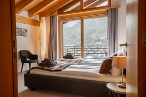 Quarto com cama, poltrona, abajur e vista para uma paisagem montanhosa.