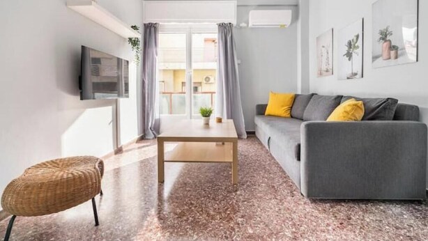 Een moderne woonkamer met een grijze bank, een houten salontafel, een rieten fauteuil en een televisie aan de muur.