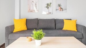Een grijze bank met gele kussens, een kleine witte tafel met een groene plant, en drie ingelijste foto's aan de muur.