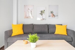 A grey sofa with yellow cushions, a small white table with a green plant, and three framed pictures on the wall. - Acropolis View by Side - Athens Central (Athina)