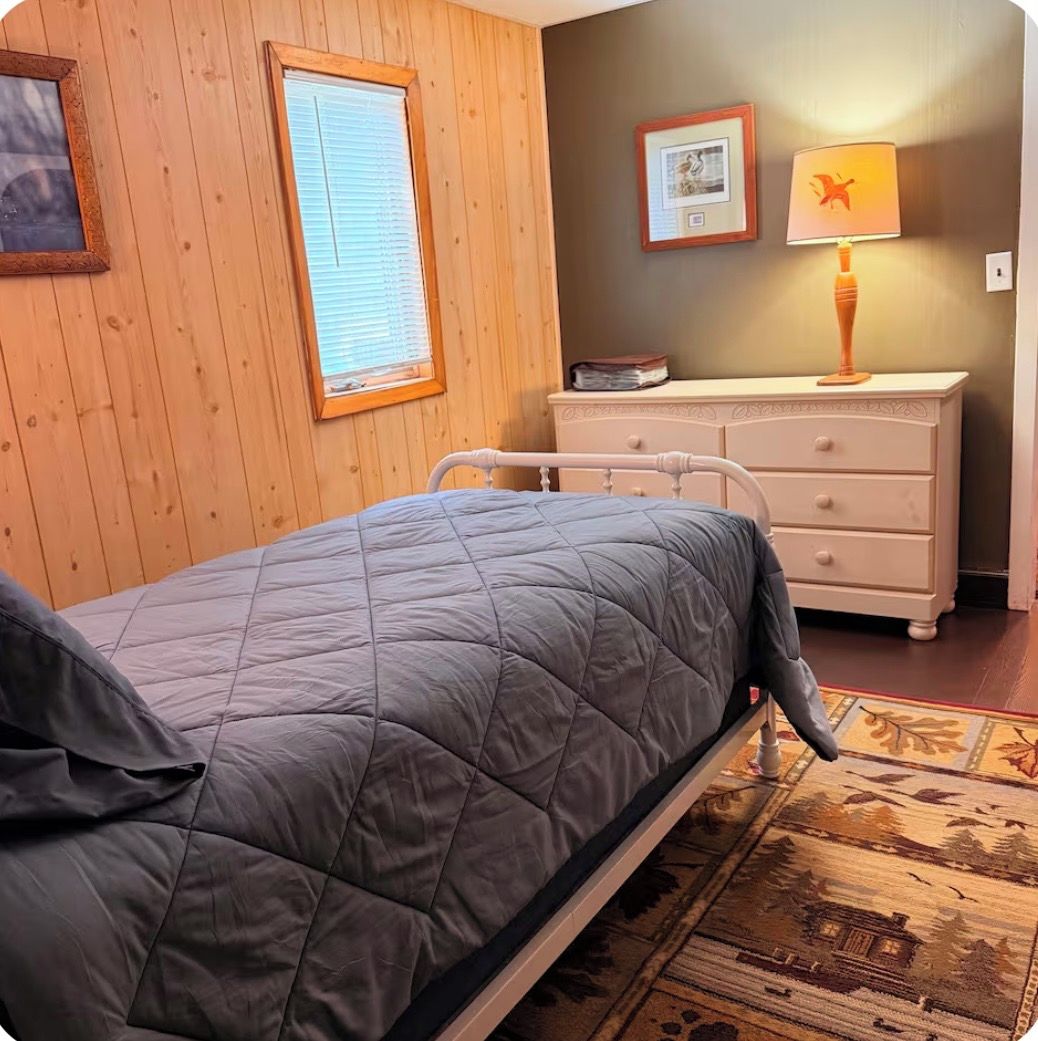 Un lit simple recouvert d’une couverture matelassée grise, une commode en bois avec des tiroirs, un tableau encadré accroché au mur et une lampe décorée d’un motif de dragon.