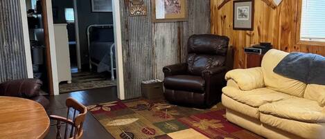 A cozy living room with a wooden floor, a leather armchair, a sofa, a dining table with chairs, and a patterned rug.