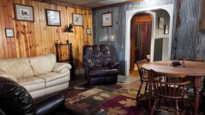 A room with a wooden wall, a leather sofa, a leather armchair, a dining table with chairs, and framed pictures on the wall.
