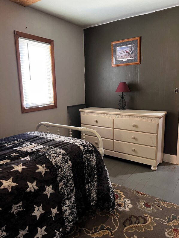 Un dormitorio con cama, cómoda, ventana y un cuadro en la pared.