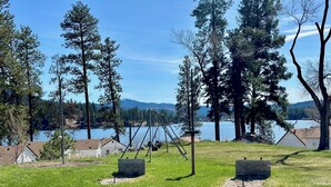 A grassy area with swings, a wooden structure, and a lake with trees in the background.