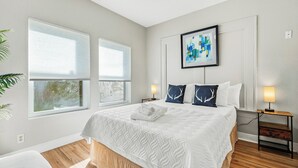 A bedroom with a large bed, two bedside tables with lamps, a framed abstract painting, and a window with blinds. - Home near NRG - Sleep 8 - WolrdCup ready (Houston)
