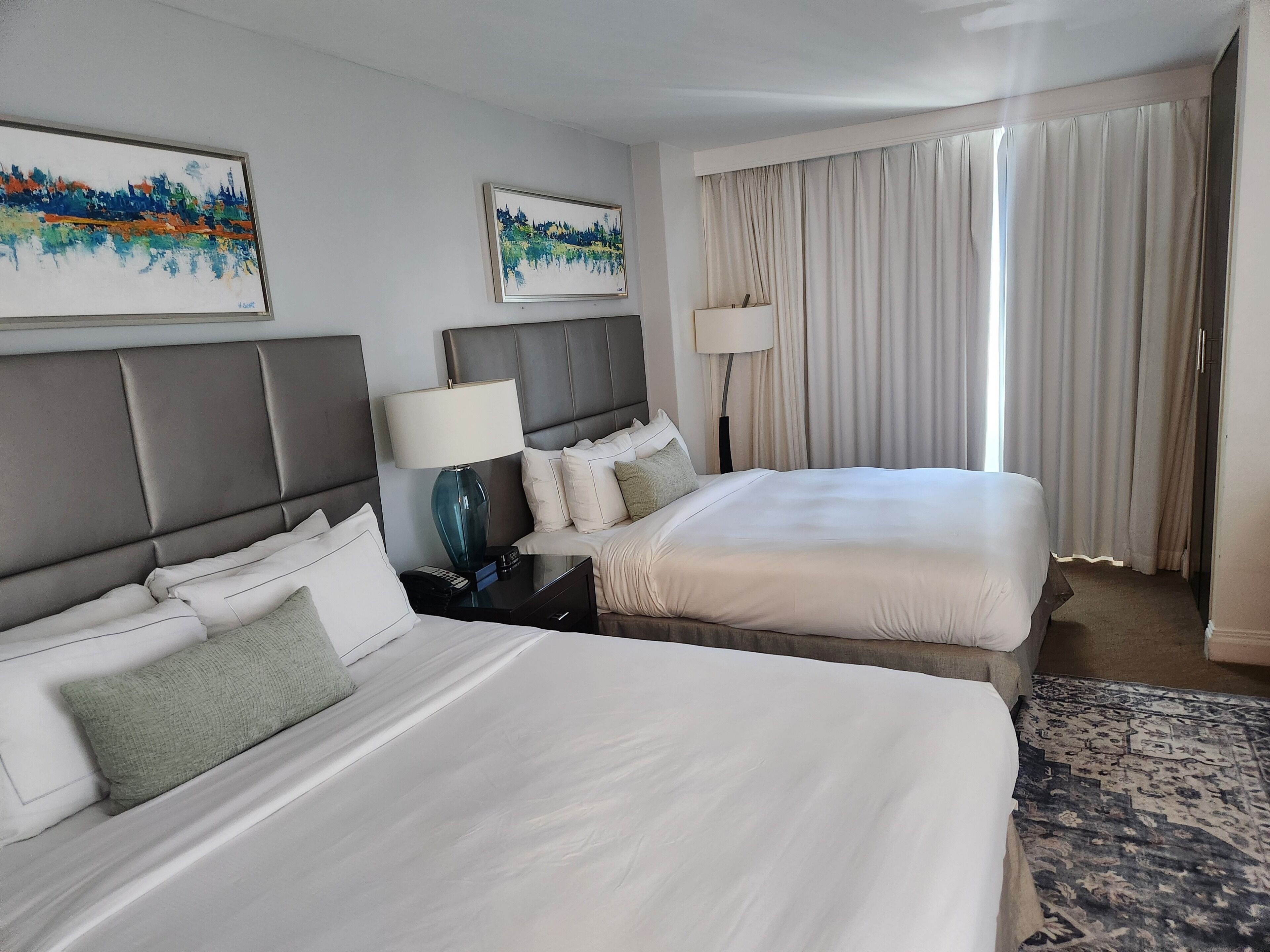 A hotel room with two beds, a nightstand with a lamp, a large window with curtains, and two paintings on the wall.