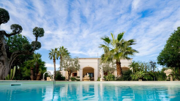 A pool with a clear blue sky, palm trees, and a building with an arched entrance. - Apartment 'Don Onofrio - Domitilla' with Mountain View, Wi-Fi and Air Conditioning (Monopoli)