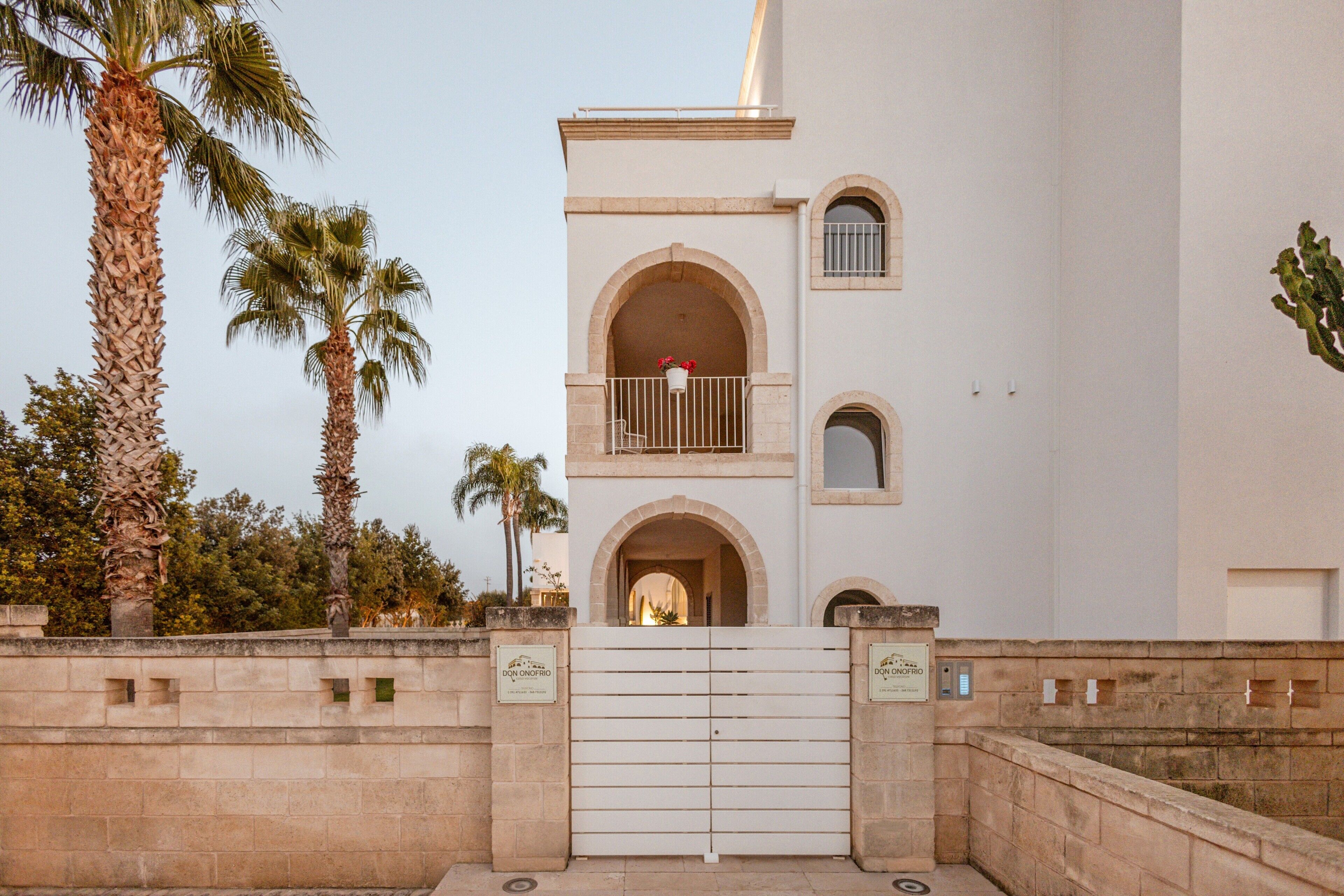 A building with a gated entrance, palm trees, and a balcony with a red flower pot.
