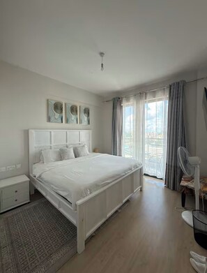 Une chambre avec un grand lit, une table de chevet, une chaise, un ventilateur et une vue sur l’extérieur.