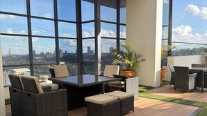 Un espacio lounge exterior moderno con un ventanal grande que da a una vista urbana, amoblado con sillas de mimbre y una mesa central.