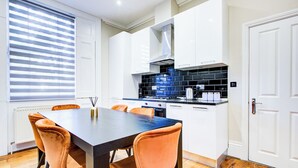 A modern kitchen with white cabinets, a black countertop, and a wooden dining table with orange chairs. - Hybrid Resi Battersea (London)