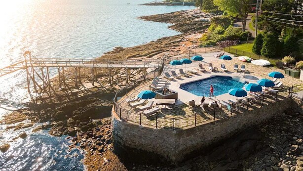 A circular pool with lounge chairs and umbrellas, surrounded by a rocky shoreline and a pier extending into the water.