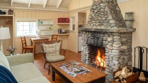 A cozy living room with a stone fireplace, wooden ceiling, and a dining area with a table and chairs. - Tanager Cottage | 3BR + 2 Porches + Fireplace (Boothbay Harbor)
