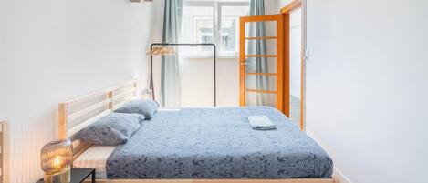 Een slaapkamer met een bed, nachtkastje, lamp, raam en gordijnen.