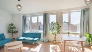 Een moderne woonkamer met een blauwe bank, een houten tafel en een eethoek met stoelen. Er zijn grote ramen met gordijnen en er staan potplanten in de kamer.