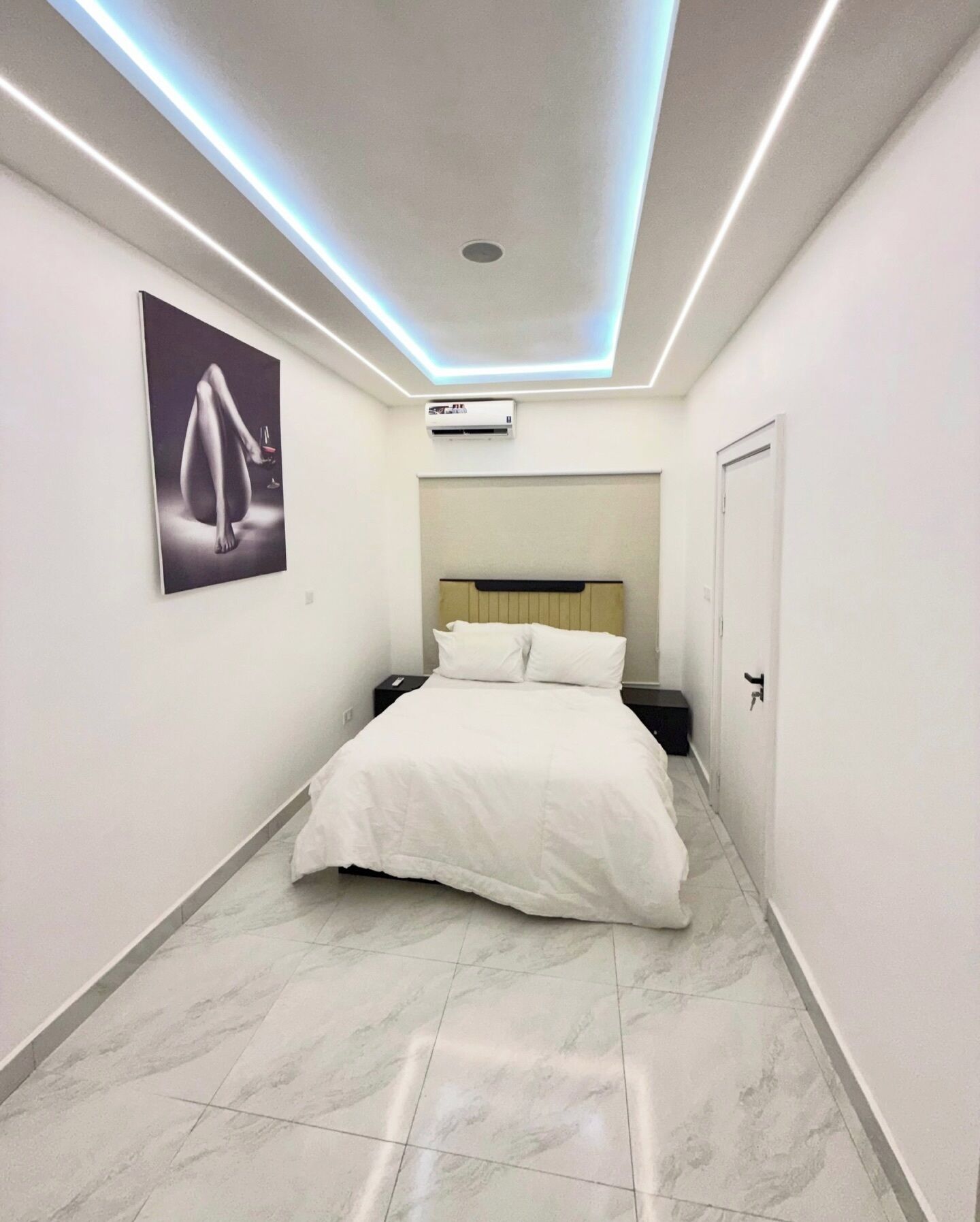 A modern hotel room with a bed, white walls, a ceiling light fixture, and a framed artwork on the wall.