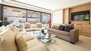 A modern living room with a sofa, armchairs, a glass coffee table, and a flat-screen TV mounted on a wooden wall. - Modern Appartement a/c 2br/4p - Cannes (Cannes)
