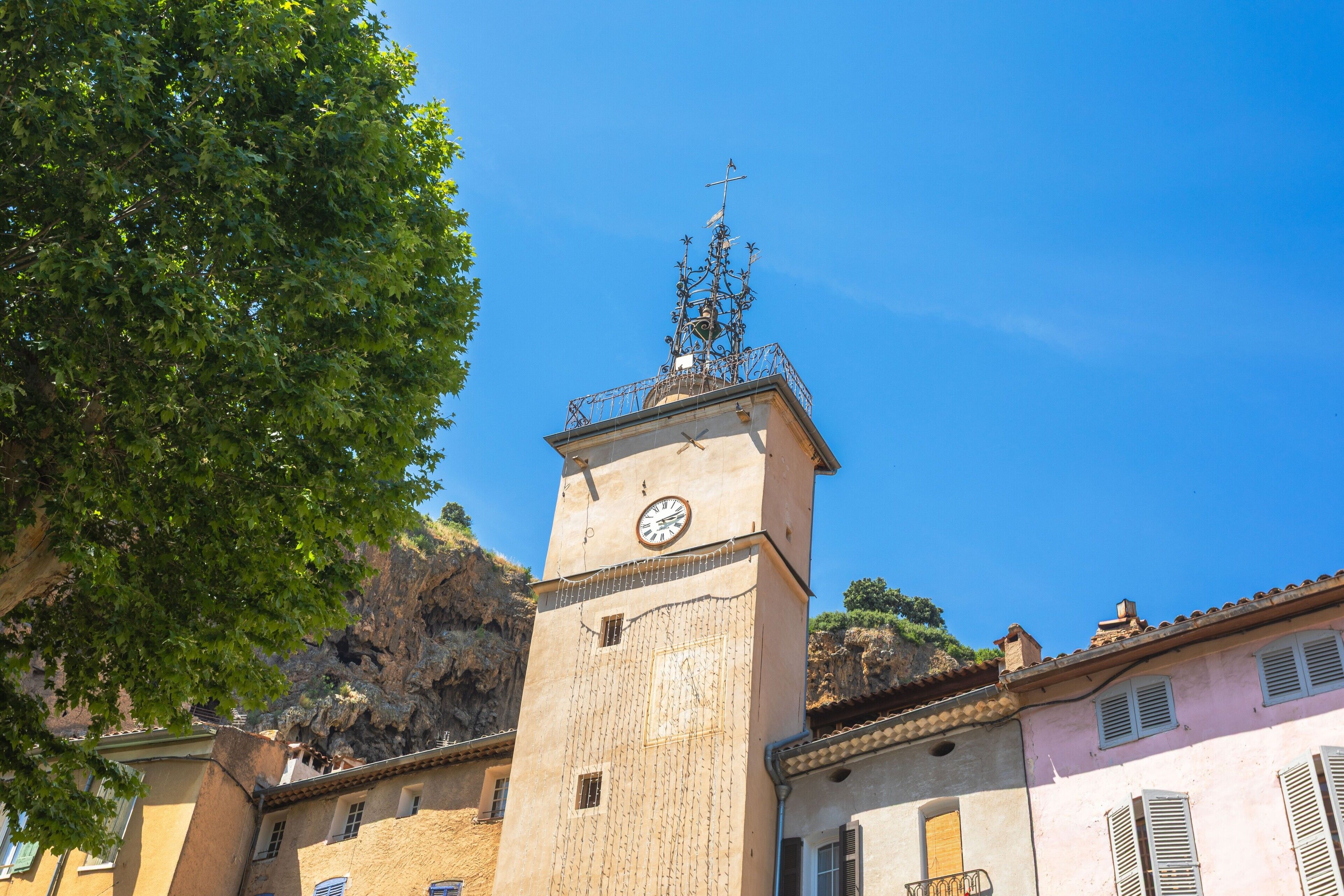 Una torre con un orologio e una croce in cima, circondata da edifici con persiane.