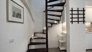 Una escalera moderna con acabado oscuro, paredes blancas y una zona de cocina con armarios blancos y estante de rejilla negra.