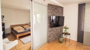 A modern bedroom with a bed, a TV mounted on a stone wall, a small table with a vase, and a sliding door leading to another room. - Modern vacation apartment in Dülmen: close to the city with a balcony view of the countryside (Dülmen)